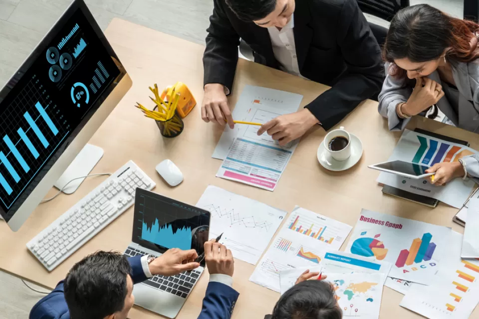 Overhead view of people at a large table working with charts and graphs on laptops and on paper